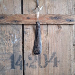 May include: A dark, metallic shoehorn hangs against a weathered wooden background. The shoehorn has a rounded end and a hole for hanging, with text and a design etched into its surface. A white string is used for hanging.