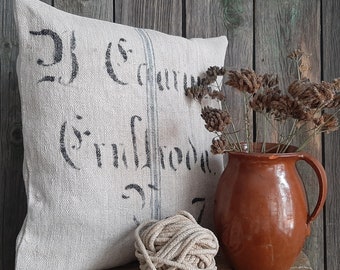 May include: A square, off-white pillow with faded black text and blue stripes. A brown ceramic pitcher holds dried flowers. A ball of beige rope sits in front of the pillow. The items are on a wooden surface.