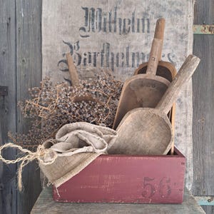 May include: A red wooden box, marked with the number "56", contains several antique wooden scoops. A burlap sack and dried berries enhance the vintage feel. A faded linen sack with text is in the background, adding to the rustic charm.