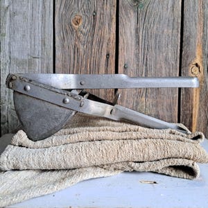 May include: A vintage, silver-toned potato ricer with a long handle and a semi-circular pressing plate. It rests on a stack of beige, textured kitchen towels. The background is a weathered wooden wall, creating a rustic aesthetic.