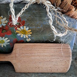 Può includere: Una spatola per burro in legno con superficie scanalata è posta davanti a un contenitore di legno dipinto. Il contenitore è decorato con fiori rossi e gialli su sfondo blu. Una corda ritorta è drappeggiata sul contenitore.