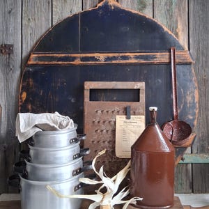 Puede incluir: Naturaleza muerta rústica con utensilios de cocina antiguos. Incluye una pila de ollas de aluminio, una jarra marrón, un rallador y un cucharón de mango largo. Una tabla de cortar de madera y una mazorca de maíz completan la escena, sobre un fondo de madera envejecida.