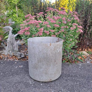 Pode incluir: Um vaso de metal cilíndrico e desgastado está sobre uma superfície de concreto. O vaso cinza tem uma textura ligeiramente áspera e está vazio. Plantas com flores rosas e vegetação são visíveis ao fundo, criando um cenário de jardim.