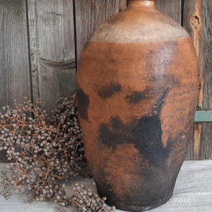 Puede incluir: Un jarrón grande de cerámica marrón con un aspecto rústico y envejecido. El jarrón tiene un cuerpo ancho y redondo y un cuello estrecho. La superficie del jarrón está cubierta de vetas marrones oscuras y negras, lo que le da un aspecto único y desgastado.