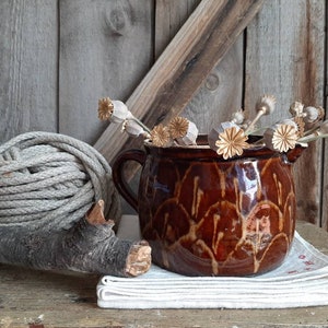 May include: A brown ceramic pitcher with a rustic design, filled with dried poppy seed heads. The pitcher is sitting on a white linen cloth, with a piece of driftwood and a ball of gray twine in the background.