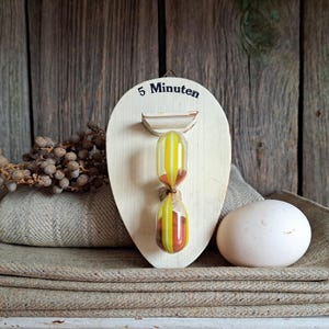 Op de afbeelding: Een houten zandloper met geel en oranje zand, gemonteerd op een houten voet. De voet is eivormig en heeft de tekst "5 Minuten" erop gedrukt.