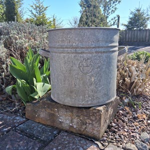 Może przedstawiać: Zniszczone, ocynkowane wiadro z dwoma uchwytami, stojące na cegle. Wiadro ma lekko zwężający się kształt i wytłoczony emblemat. Tło przedstawia zieleń i czyste, błękitne niebo.