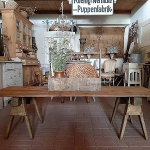 Könnte beinhalten: Ein rustikaler Holztisch mit zwei Bockbeinen. Der Tisch ist aus Naturholz gefertigt und hat eine gewitterte Oberfläche. Der Tisch steht in einem Raum mit einer Vielzahl von antiken Möbeln und Dekorationen.