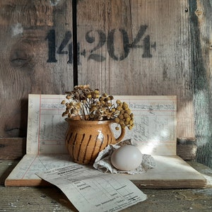 Può includere: Un'anfora di ceramica marrone con un uovo bianco e fiori secchi su un libro mastro vintage con il numero 14204 scritto su uno sfondo di legno.