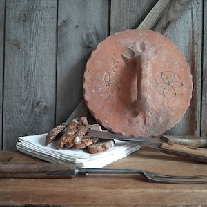 Könnte beinhalten: Ein rustikaler Holztisch mit einer Brotkuppel aus Terrakotta, einem Stapel geschnittenem Brot, einem Messer mit Holzgriff und einer Gabel mit Holzgriff. Die Kuppel hat einen Griff und dekorative Designs.