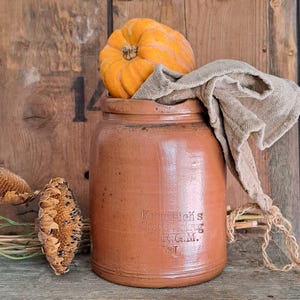 May include: A rustic, brown stoneware crock with the text "Krumrich's Conservenkrung D.R.G.M. 3L" is filled with a small orange pumpkin and a tan cloth. The crock sits on a wooden surface with dried sunflowers.