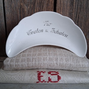 Könnte beinhalten: Eine weiße Porzellanschale mit gewelltem Rand und schwarzem Text "Für Braten u. Schalen" liegt auf zwei gefalteten Leinentüchern. Die Tücher sind beige und cremefarben, mit einem roten Stickmuster auf dem unteren Tuch. Der Hintergrund ist eine rustikale Holzoberfläche.