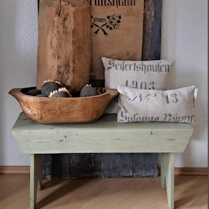 May include: A distressed green wooden bench supports a wooden bowl with decorative items. Two pillows with printed text and a large wooden log are behind the bench. The scene evokes a vintage aesthetic, with a focus on natural materials.