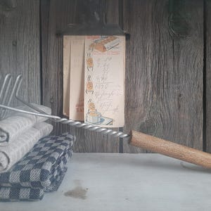 May include: A silver potato masher with a wooden handle rests on top of a stack of folded gray and black checkered fabric towels. A vintage handwritten recipe card is attached to a wooden wall behind the masher.