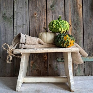 Puede incluir: Naturaleza muerta rústica con tres calabazas sobre un banco blanco desgastado y un paño beige. Las calabazas son verdes, blancas y naranjas, con superficies texturizadas. El fondo es una pared de madera envejecida, creando un tema otoñal.
