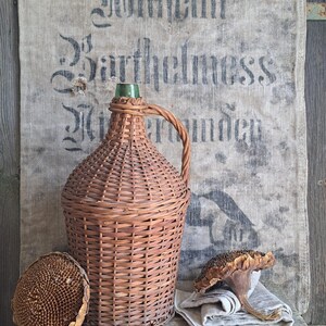 May include: A brown wicker-covered bottle with a green glass top and a woven handle. The bottle sits on a wooden surface with a dried sunflower head and a linen cloth. A burlap sack with faded text is in the background.