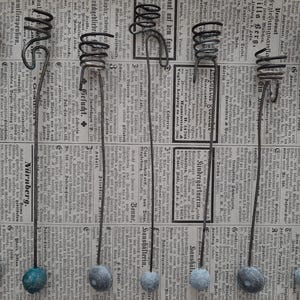 May include: Six handmade metal bottle stoppers with a coiled top and a long stem, each ending with a small, round, painted bead. The stoppers are displayed against a backdrop of vintage newspaper.