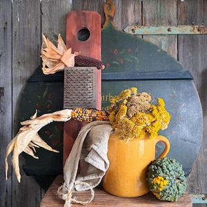 May include: A rustic still life featuring a red cutting board, a dark green painted wooden shape, and a yellow ceramic pitcher filled with dried yellow flowers.  Ears of corn, a grater, and a tan cloth complete the arrangement. A green and yellow gourd sits next to the pitcher.