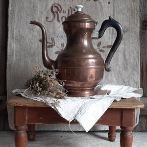 May include: A copper coffee pot with a black handle and a white knob on the lid. The pot is sitting on a wooden stool with a white cloth draped over it.