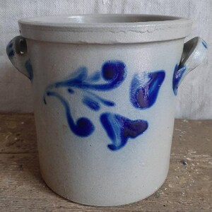 May include: A white ceramic crock with blue floral designs and two handles. The crock is a traditional style with a rustic look.