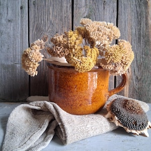 May include: A rustic still life featuring a brown ceramic pot filled with dried yellow flowers. A tan linen cloth and a dried sunflower head rest on a weathered wooden surface. The background is a wooden wall.