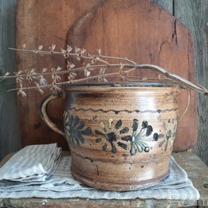 May include: A brown ceramic pot with a floral design in black, green, and yellow. The pot has a handle and is sitting on a wooden surface.