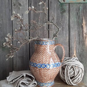 Könnte beinhalten: Eine braune Keramikkanne mit blauen und weißen Blumenmustern, gefüllt mit getrockneten Zweigen, steht auf einer Holzoberfläche mit einer Schnurkugel.