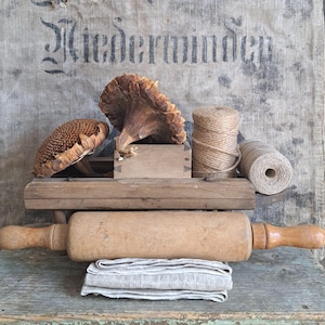 Può includere: Una natura morta rustica con un mattarello di legno, rocchetti di spago e teste di girasole essiccate. Un panno di lino piegato è posto sotto il mattarello. Lo sfondo include un sacco di iuta con testo sbiadito.