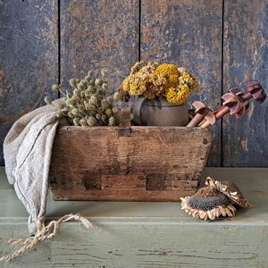 Könnte beinhalten: Eine rustikale Holzkiste gefüllt mit getrockneten Blumen, einem Leinentuch und dekorativen Holzobjekten. Die Kiste steht auf einer grünen Oberfläche vor einem verwitterten Holz-Hintergrund. Die Anordnung umfasst gelbe und grüne florale Elemente.