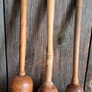 May include: Three antique wooden muddlers with varying shapes and sizes. The muddlers are made of light and dark brown wood, with cylindrical and rounded handles. They are displayed against a rustic wooden background.