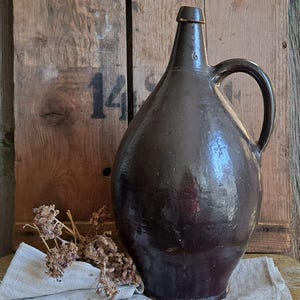 May include: A large, dark brown antique jug with a handle. The jug has a rounded body and a narrow neck. It sits on a gray linen cloth, with dried flowers and a wooden background. The number "14" is visible on the wooden background.