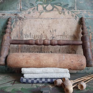 May include: A vintage wooden clothes wringer with a large wooden roller and two ornate handles. Below the wringer are three folded linen towels in shades of grey and blue. Dried seed pods are in the foreground.