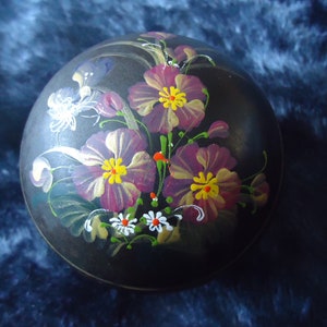 Op de afbeelding: Een zwarte ronde doos met een geschilderd bloemenontwerp. De bloemen zijn paars, geel en wit.