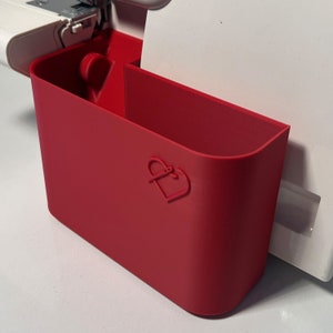 Könnte beinhalten: Ein roter Plastik-Nähmaschinen-Zubehör-Organizer mit einem herzförmigen Logo. Der Organizer hat zwei Fächer und ist so konzipiert, dass er an der Seite einer Nähmaschine befestigt werden kann.