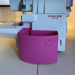 Może przedstawiać: Biała maszyna do szycia Singer Ultra Lock z jasnoróżowym pojemnikiem na odpady. Pojemnik ma kształt serca. Maszyna stoi na białej powierzchni.