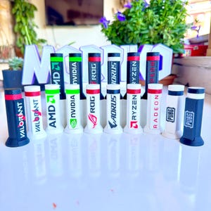 Könnte beinhalten: Eine Sammlung zylindrischer Gaming-Accessoires. Die Accessoires zeigen verschiedene Logos und Texte, darunter "VALORANT", "AMD", "NVIDIA", "ROG", "AORUS", "RYZEN", "RADEON" und "PUBG". Das Farbschema umfasst schwarze, weiße, rote und grüne Akzente.