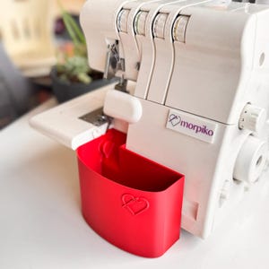 May include: A white Morpiko serger sewing machine featuring a bright red plastic scrap catcher bin attached to the front. The bin has a heart-shaped logo with a needle icon embossed on the center. The machine has multiple tension dials on top.