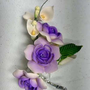 May include: A floral arrangement featuring handcrafted flowers in shades of purple and white. The arrangement includes calla lilies, roses, and green leaves. The flowers are attached to a decorative silver stem, perfect for a wedding or special event.
