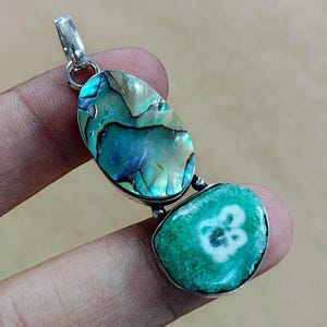 Peut inclure: Un pendentif composé de deux pierres polies. La pierre supérieure est ovale avec des reflets bleus et verts irisés. La pierre inférieure est ronde, vert vif avec un motif blanc. Le pendentif a un bélière argenté.