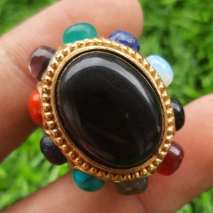 May include: A gold-toned ring featuring a large, oval black stone at the center, surrounded by an array of colorful, small gemstones. The ring's design includes red, green, blue, and other colored stones, creating a vibrant and eye-catching piece of jewelry.