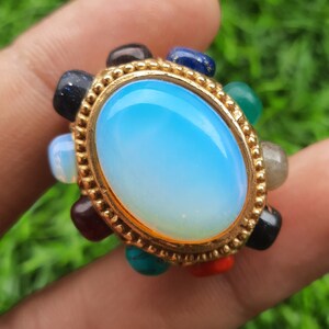 May include: A gold ring with a large oval-shaped blue opal gemstone surrounded by smaller gemstones in various colors, including red, green, black, and white.