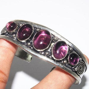 May include: Silver cuff bracelet with five oval-shaped purple gemstones. The bracelet has a decorative design with swirls and small beads.