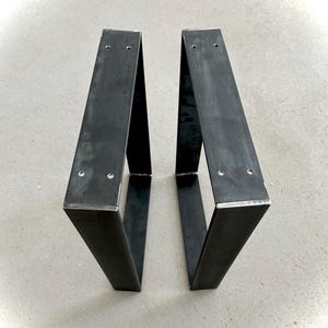 Könnte beinhalten: Zwei dunkle Metall-Tischbeine mit V-förmigem Design. Jedes Bein hat eine rechteckige Oberseite mit vorgebohrten Löchern zur Montage. Die Beine bestehen aus dickem, robustem Metall und haben einen rohen, industriellen Look. Die Beine sind für eine Tischplatte konzipiert.