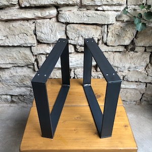 Könnte beinhalten: Zwei schwarze Metalltischbeine mit quadratischer Form und geschweißter Konstruktion. Die Beine stehen auf einer Holzoberfläche.