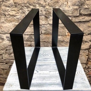Könnte beinhalten: Zwei schwarze Metall-Tischbeine mit geometrischem Design. Die Beine sind abgewinkelt und haben eine rechteckige Form. Sie stehen auf einer weißen Holzoberfläche vor einer Steinmauer.