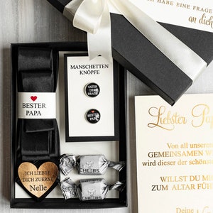 May include: A black gift box with a white satin ribbon and bow. The box contains a black bow tie, cufflinks, and a heart-shaped wooden plaque with the text "I loved you first, Nelle". The box also includes a card with the text "Dearest Dad, From all our shared journeys, this will be the most beautiful. Will you walk me down the aisle? Your Nelle, 25.05.2025".