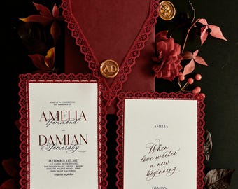 Könnte beinhalten: Ein burgunderroter Umschlag mit Spitzenrand und goldenem Wachssiegel mit den Buchstaben AD, begleitet von zwei weißen Hochzeitseinladungskarten mit burgunderroten Spitzenrändern, schwarzem Text und floralen Elementen auf schwarzem Grund.