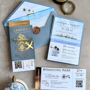 Puede incluir: Un conjunto de invitaciones de boda con temática de viaje que incluye un folleto azul estilo pasaporte con texto en lámina dorada, una tarjeta de embarque con código QR y un sobre con una ilustración de isla tropical. Incluye brújula y globo.
