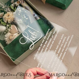 May include: A clear acrylic box with a green velvet interior and a gold heart charm. The box contains dried flowers and a clear acrylic card with the text "Lieb!" and a wedding-related message in German.