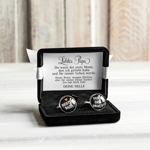 May include: A black gift box with a white satin ribbon and a card that reads "Liebster Papa Du warst der erste Mann, den ich geliebt habe und f&uuml;r immer lieben werde. Heute Braut, morgen Ehefrau, aber f&uuml;r immer Deine Tochter. Ich hab Dich lieb! DEINE NELLE". The box contains two cufflinks, one with the text "BESTE PAPA" and the other with the text "VATE DER BRAUT".
